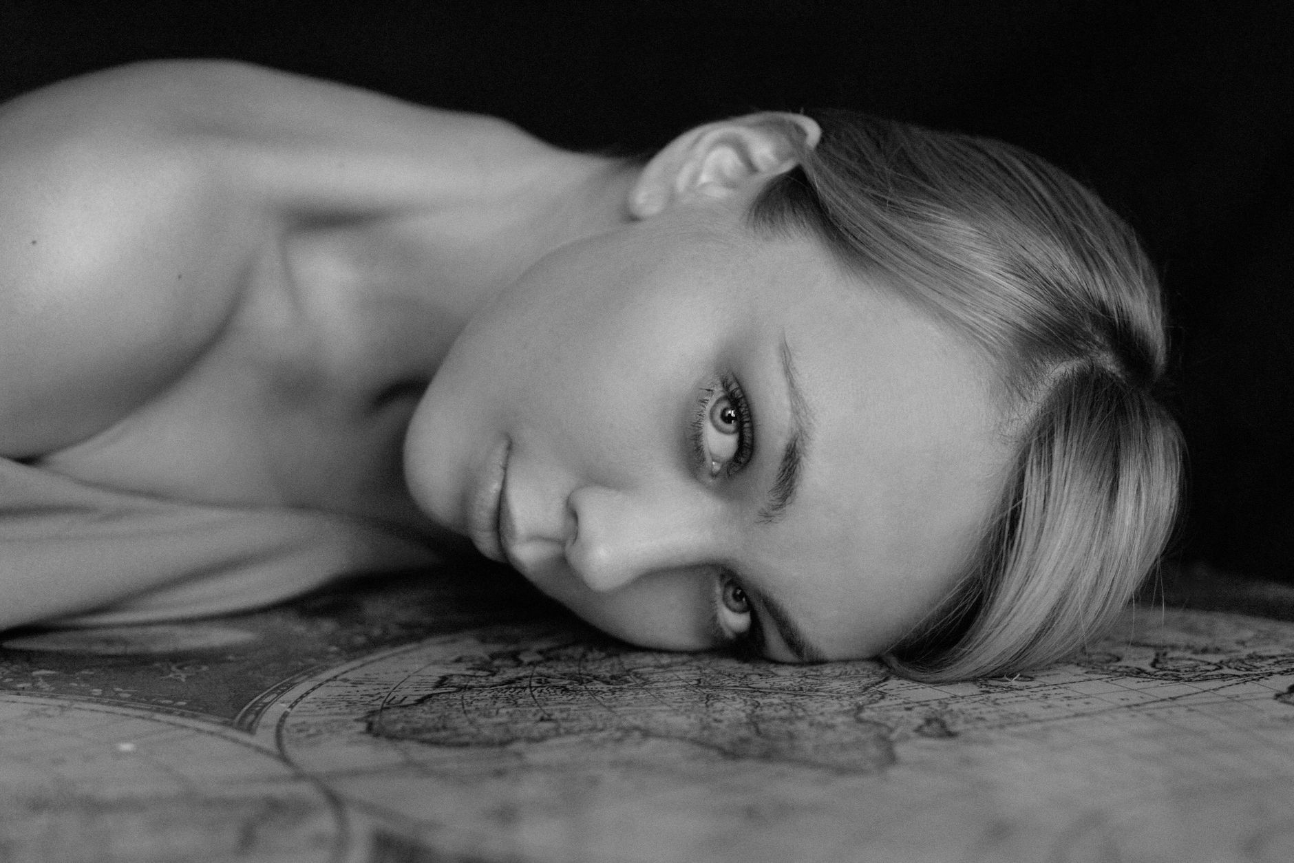 Black and white of tranquil young female lying on map table and looking at camera against dark background