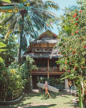 冲浪女孩拿着冲浪板，走在热带绿树环绕的平房旁