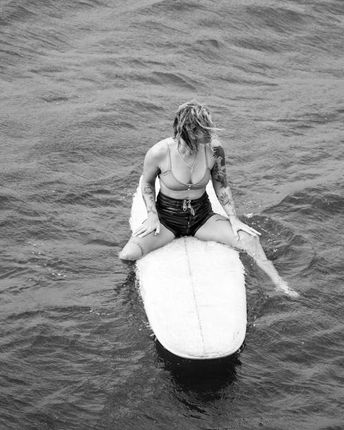 Woman in Black and White Bikini Sitting on White Surfboard on Water