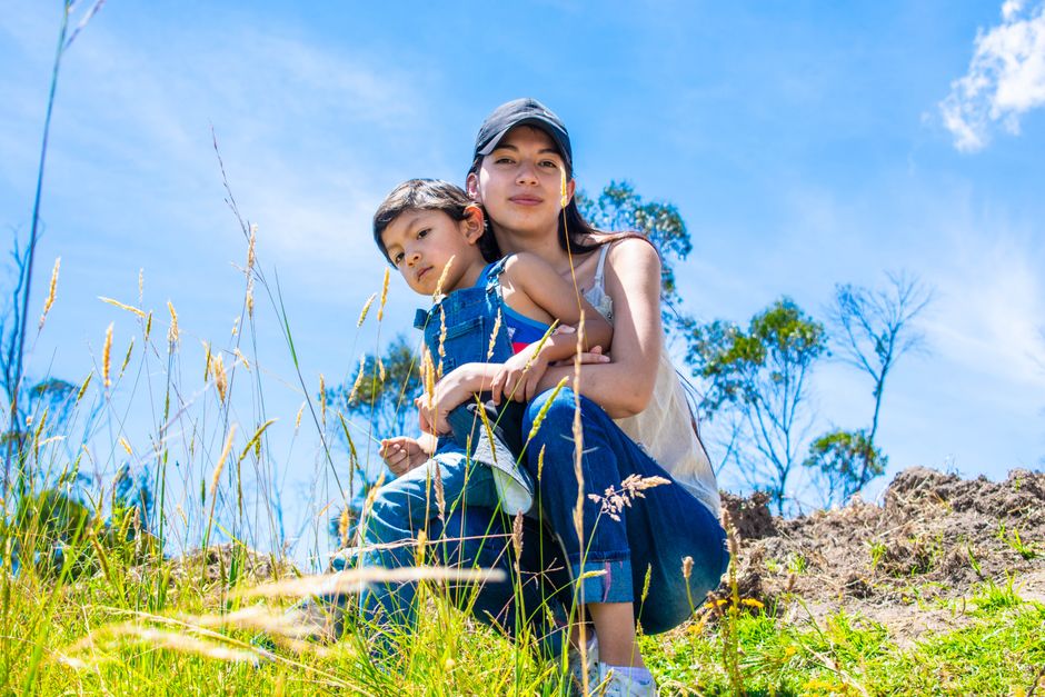 Una madre y su hijo disfrutando de un día soleado al aire libre, rodeados de hierba silvestre y cielos despejados.