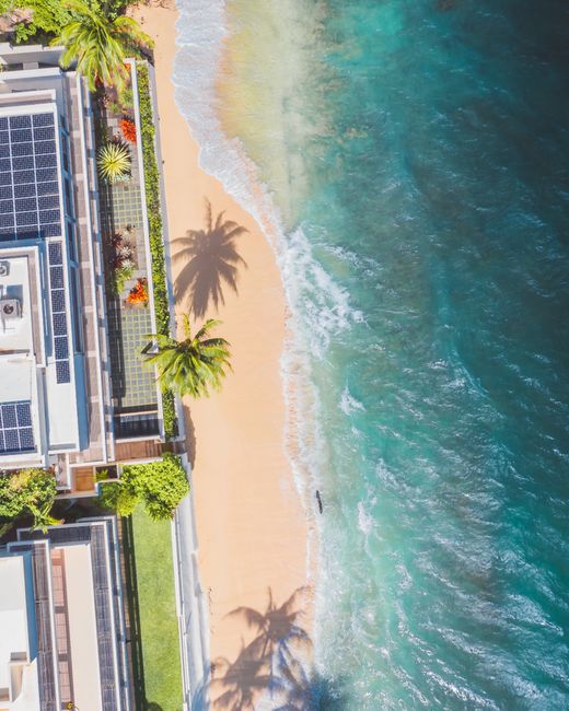 Photo by Jess Loiterton Aerial shot of a solar-powered building by a tropical beach with lush greenery and crashing waves.