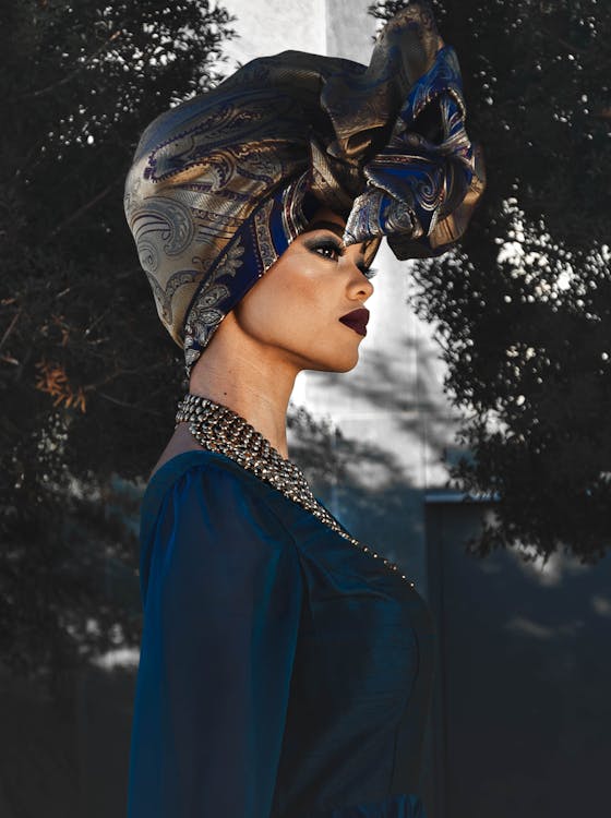 Side view of fashionable young ethnic female with makeup in stylish dress and turban standing on street near green trees and looking away