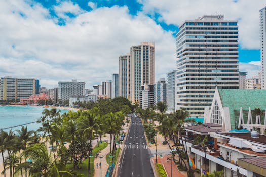 Photo by Jess Loiterton Aerial view of Waikiki's beachfront with skyscrapers and palm trees under a bright sky.