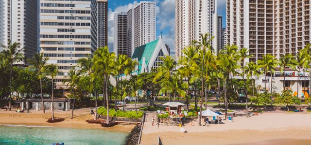Explore the vibrant Honolulu coastline with skyscrapers, palm trees, and a sandy beach under a sunny sky.