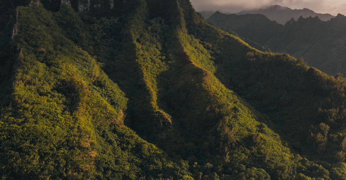 Photo by Jess Loiterton Aerial view of a lush green mountain by the sea under a dramatic, cloudy sky.