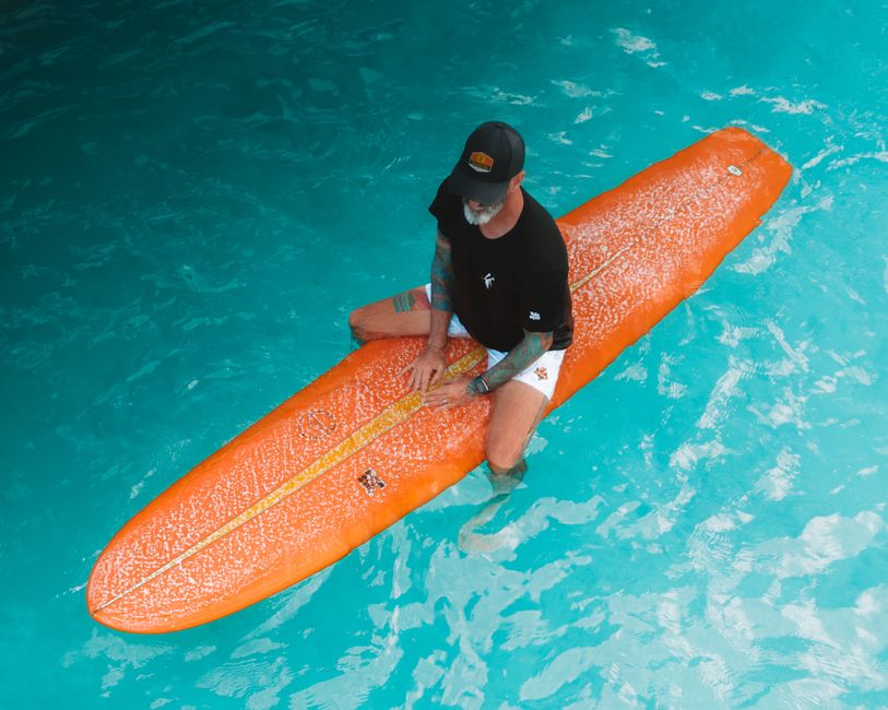 Man sitting on an orange surfboard in clear blue water, enjoying a summer day.