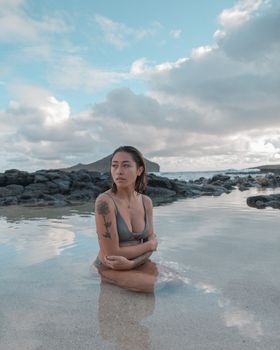 Tattooed woman in swimwear posing on a tranquil rocky beach during the day.