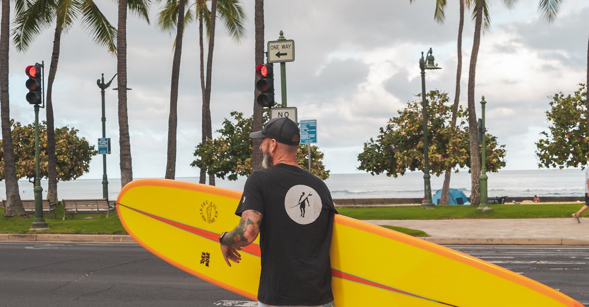 Photo by Jess Loiterton Man carrying surfboard across tropical street with palm trees overhead.