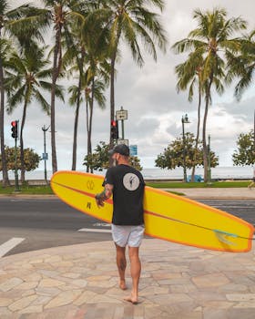 Photo by Jess Loiterton Man carrying surfboard across tropical street with palm trees overhead.