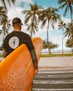 一名男子拿着冲浪板，走在热带海滩的人行道上，四周环绕着棕榈树