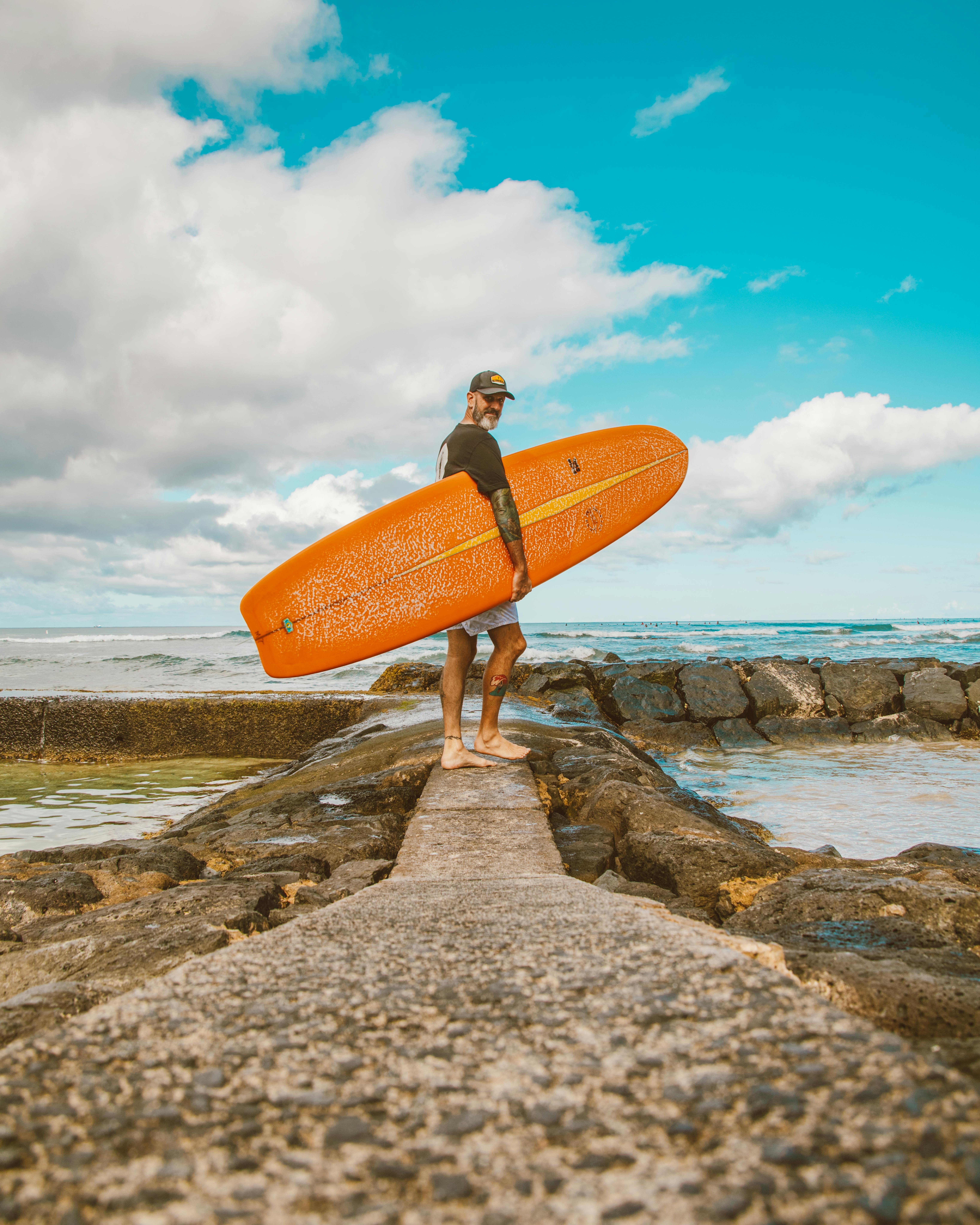 一名男子拿着冲浪板，站在海边岩石码头上，天空明亮