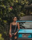 一名年轻女子拿着相机，站在一辆老爷车旁，周围是郁郁葱葱的户外环境