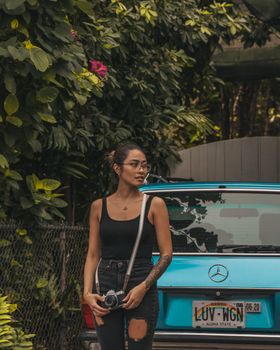 一名年轻女子拿着相机，站在一辆老爷车旁，周围是郁郁葱葱的户外环境