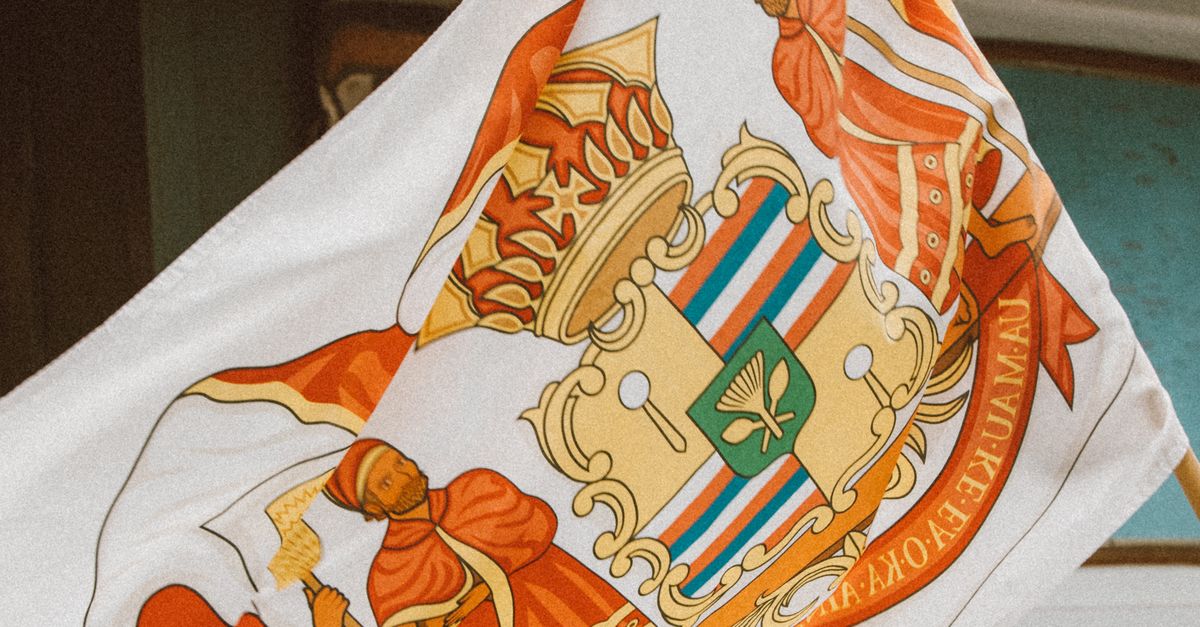 Photo by Jess Loiterton A Hawaiian flag with a distinctive coat of arms waves outdoors, symbolizing heritage and pride.