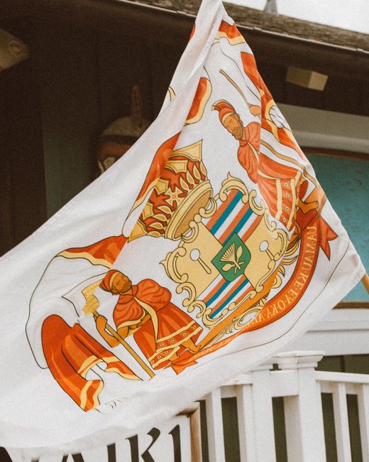 Photo by Jess Loiterton A Hawaiian flag with a distinctive coat of arms waves outdoors, symbolizing heritage and pride.