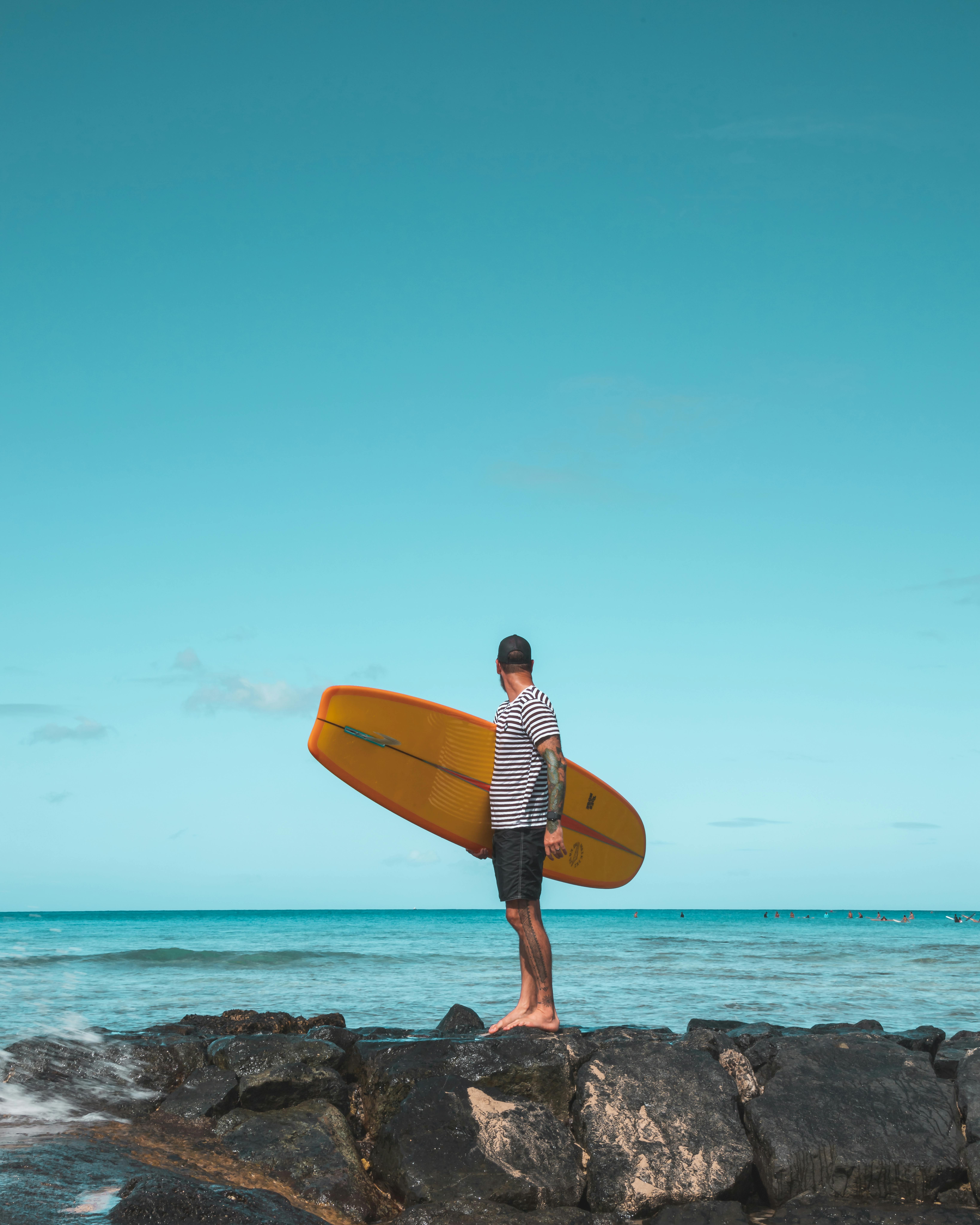 蔚蓝的天空下，一名冲浪者站在岩石海岸上，手里拿着黄色冲浪板