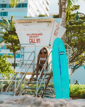 A woman sitting at a lifeguard tower with a surfboard on a sunny beach day.