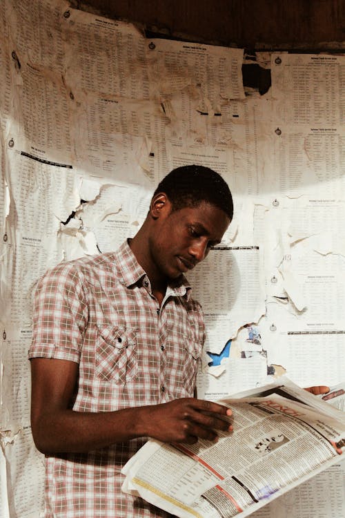 Side view of concentrated African American male with short hair in shirt reading newspaper while standing near shabby wall with torn papers in daylight