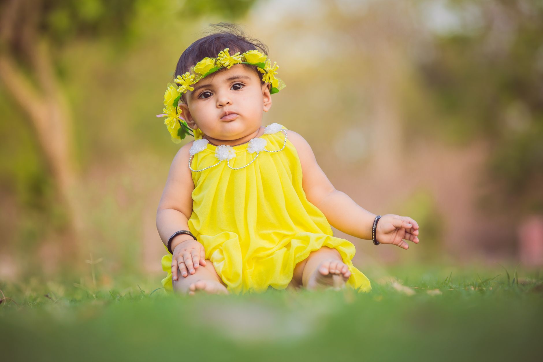 Cute baby girl wearing a yellow dress and floral headband, sitting on lush green grass outdoors.