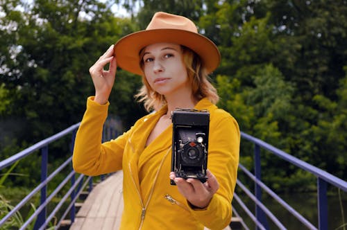 Young stylish female photographer looking at camera while standing on bridge with retro photo camera