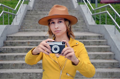 Calm female photographer in trendy outfit looking at camera while standing on street and taking photo on retro camera