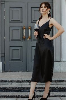 Confident young lady in black elegant dress standing near doors of aged house with hand on waist and drinking glass of wine