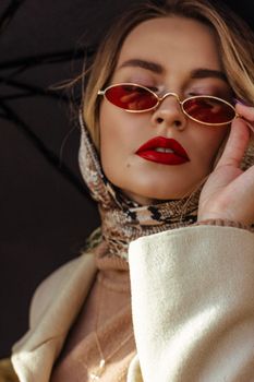 一位涂着红唇的美丽年轻女士，身着优雅服装，调整着时尚太阳镜，撑着伞站在街上，从低角度拍摄