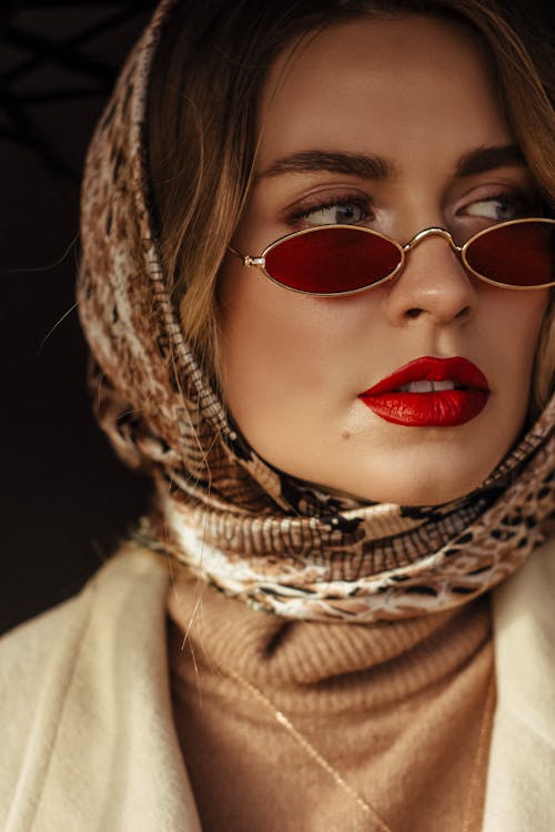 Confident young stylish lady with red lips in fashionable outfit and sunglasses standing on street on sunny day and looking away