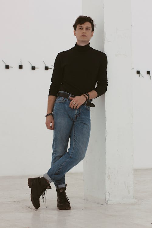 Full body of self assured young male in trendy outfit and boots leaning on white wall and looking at camera