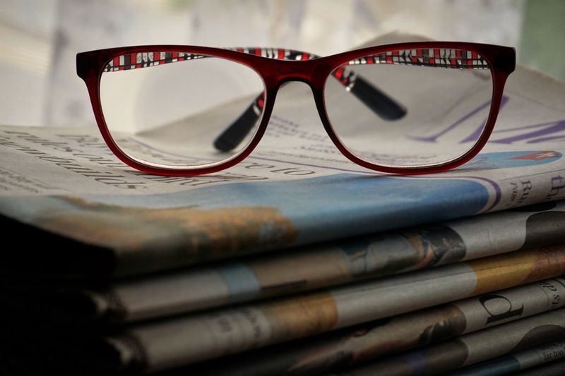 Close-up of red glasses resting on stacked newspapers, emphasizing reading and vision.