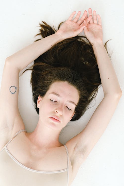 From above of young relaxed young female with long dark hair lying on white bed with closed eyes and raised arms