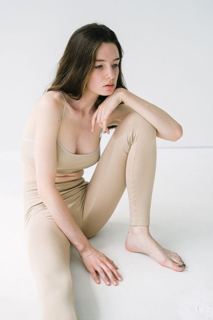 Pensive barefoot female with dark hair wearing beige top and leggings while sitting on floor with bent leg on white background