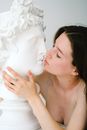 Side view of gentle female with closed eyes and dark hair kissing white plaster bust sculpture of man on white background in studio