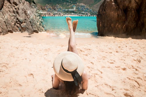 Back view of anonymous female traveler in straw hat with raised naked legs lying on sandy beach near azure sea amidst rocks