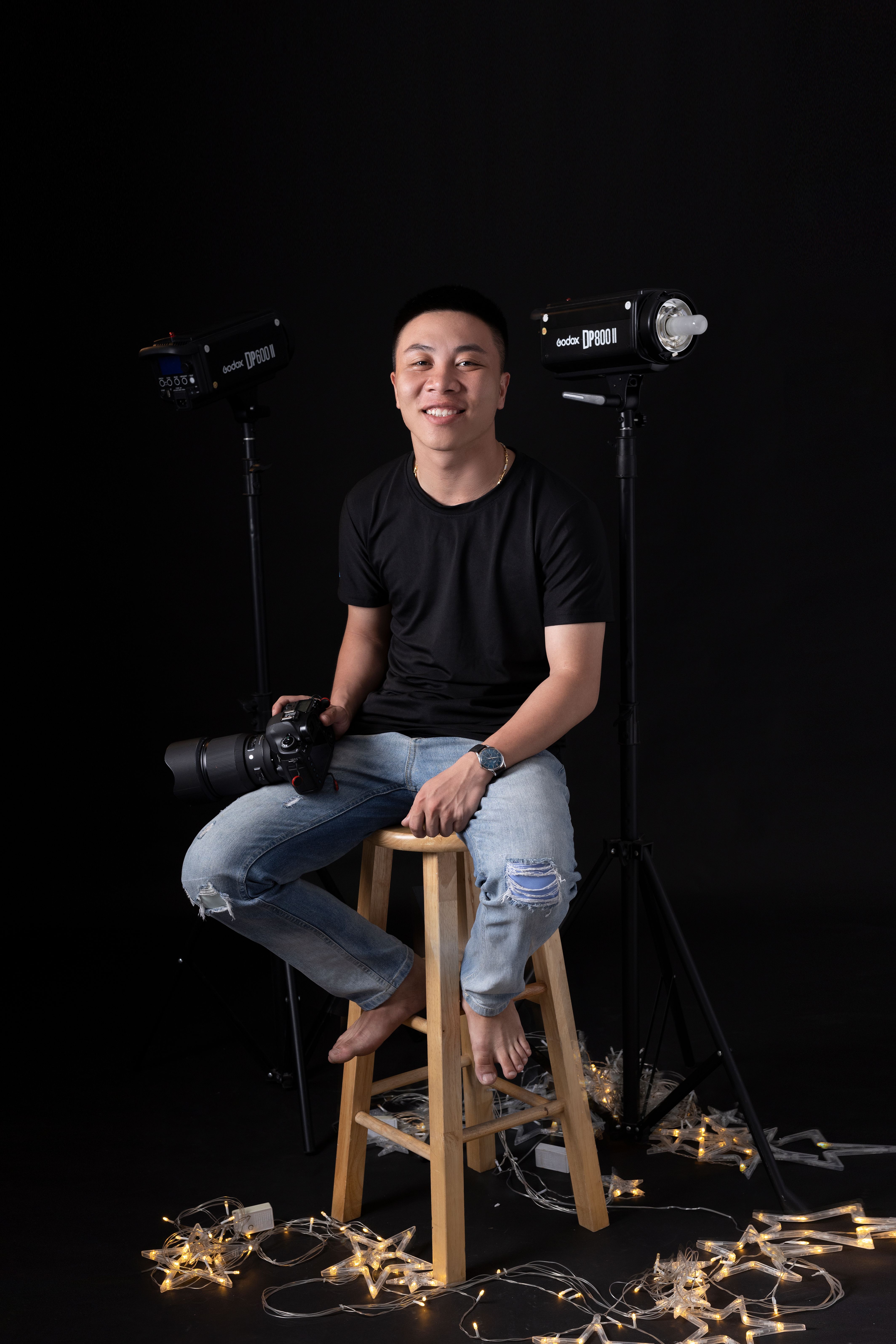 Full body of ethnic male photographer in jeans sitting on stool in studio with decoration on floor on black background while smiling and looking at camera