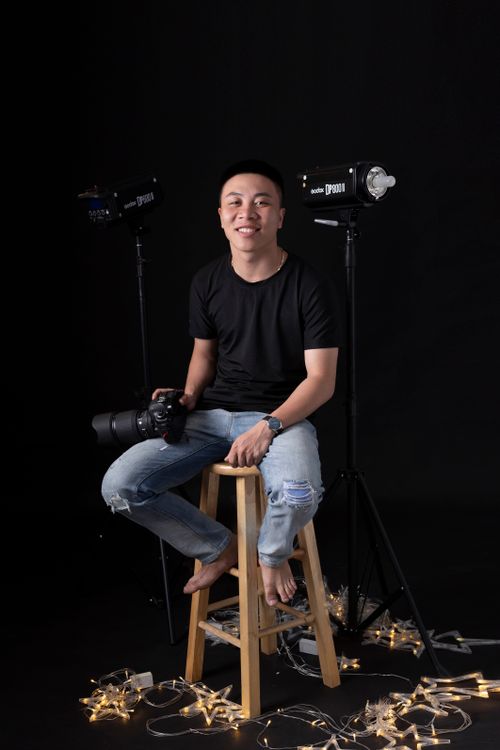 Free Full body of ethnic male photographer in jeans sitting on stool in studio with decoration on floor on black background while smiling and looking at camera Stock Photo