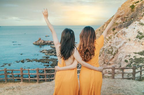 Free Back view of anonymous female friends in yellow dresses cuddling on rocky cliff while standing with raised arms near calm ocean in summer day Stock Photo