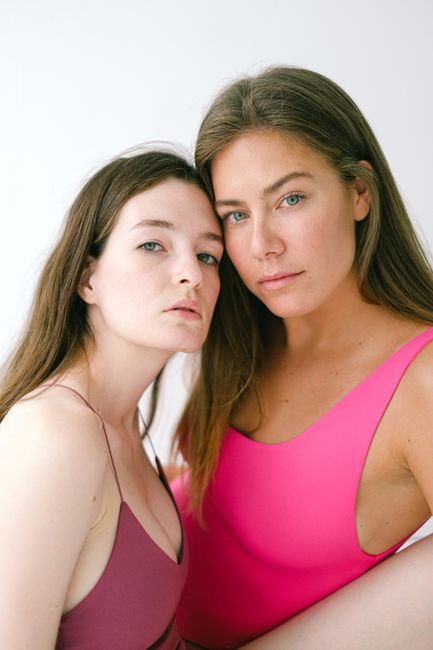 Serious female models with long hair wearing bodysuits and looking at camera while sitting on white background in light studio