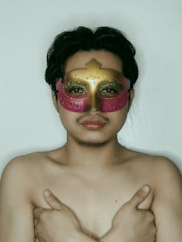 A shirtless man wearing a decorative mask poses with crossed arms on a white background.