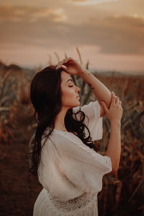Free A serene woman posing with her hand on her forehead in a field at sunset. Stock Photo