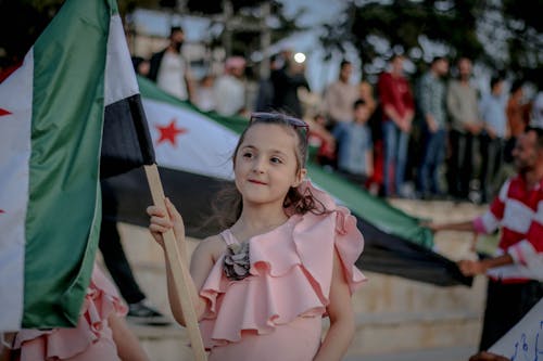 Happy girl in light pink blouse showing unofficial Syrian flag and looking away with smile on protest