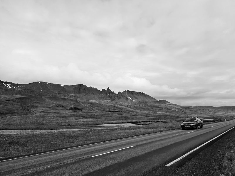 Black and white of modern automobile driving along asphalt highway between rocky mountains on cloudy day