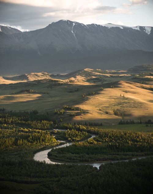 Free A breathtaking aerial view of a winding river through a lush valley with majestic mountains in the backdrop. Stock Photo