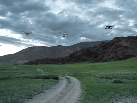 Three drones hover over a picturesque mountain landscape with a cloudy sky and winding trail.