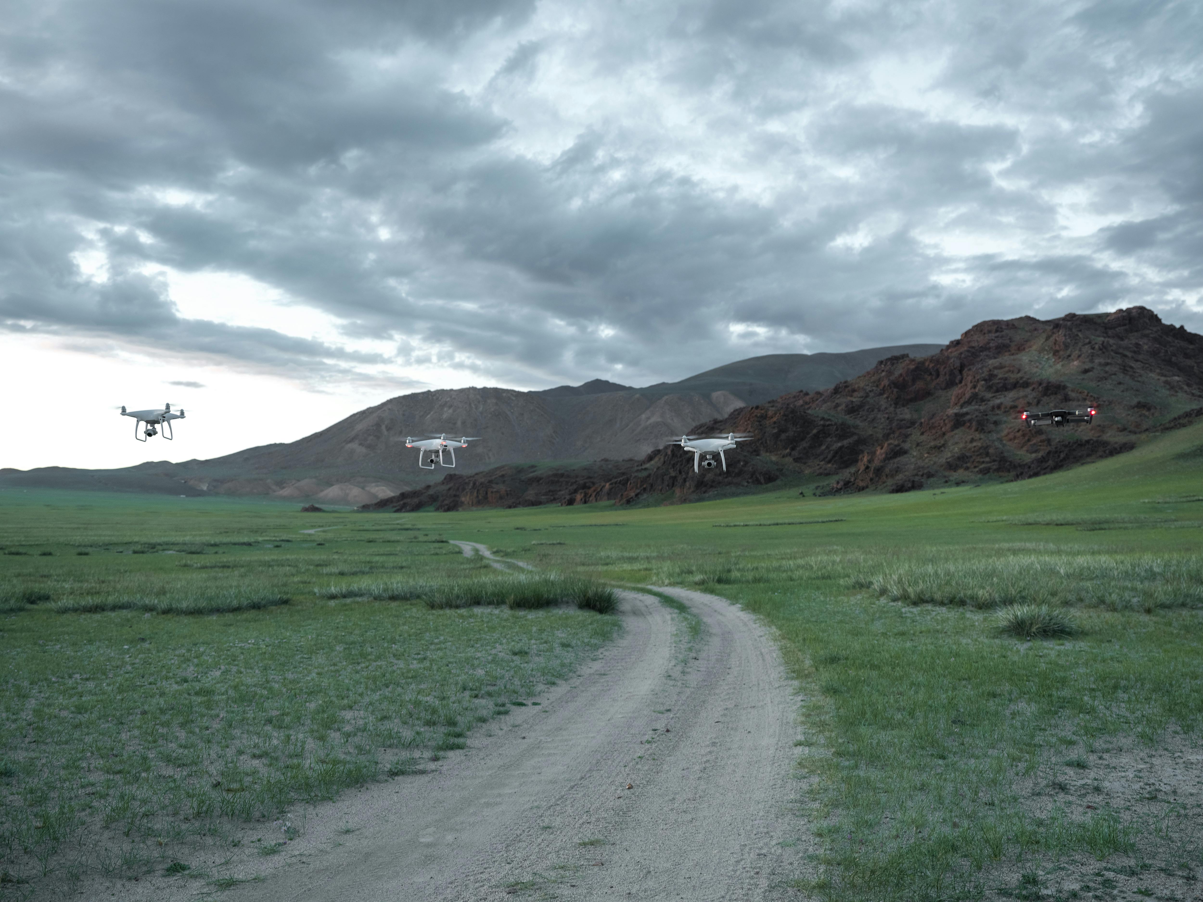 Aerial drones maneuver over a green field with dramatic clouds and rugged mountains in the background.