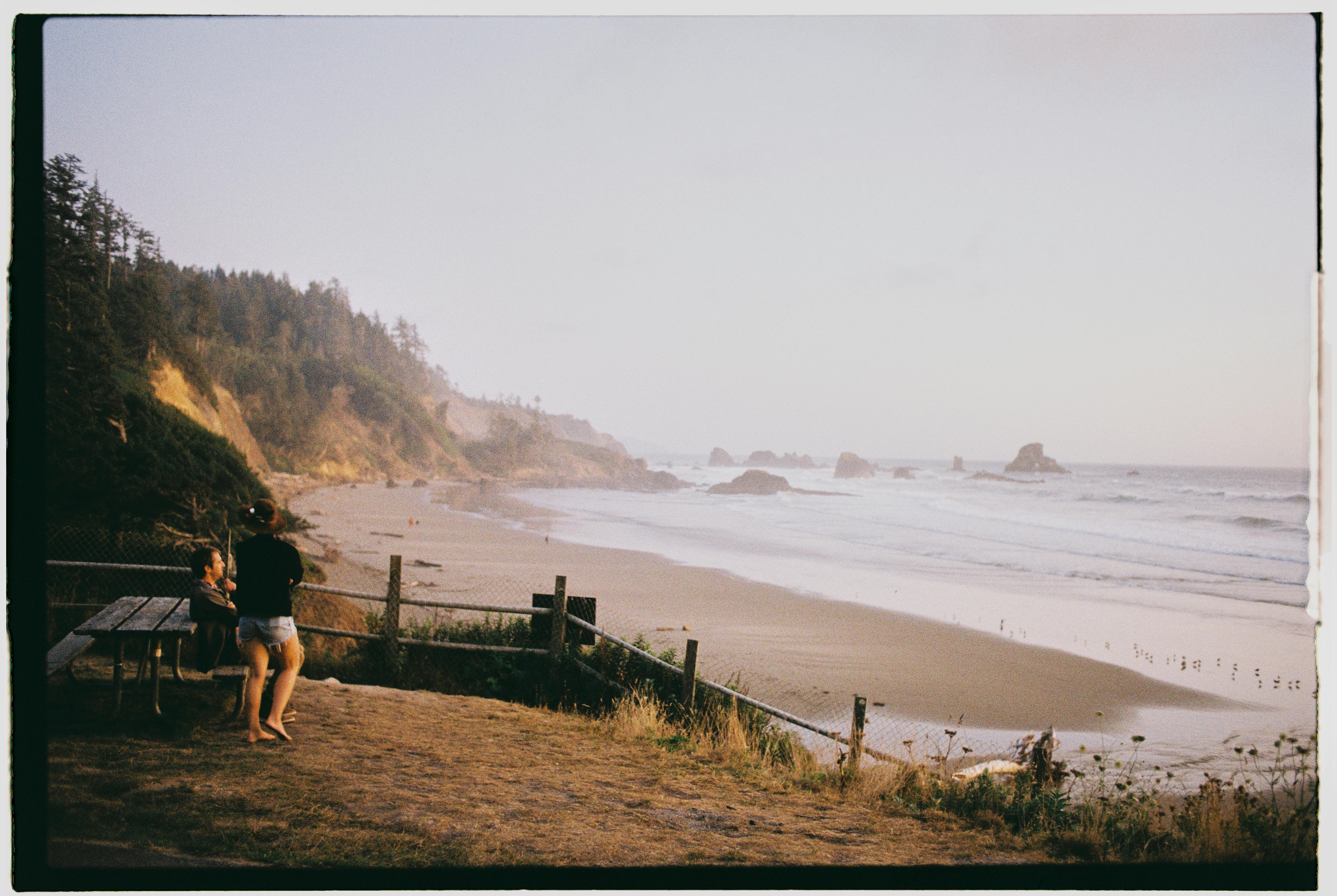 A serene beach with waves, cliffs, and people by a picnic table, captured in a warm light.