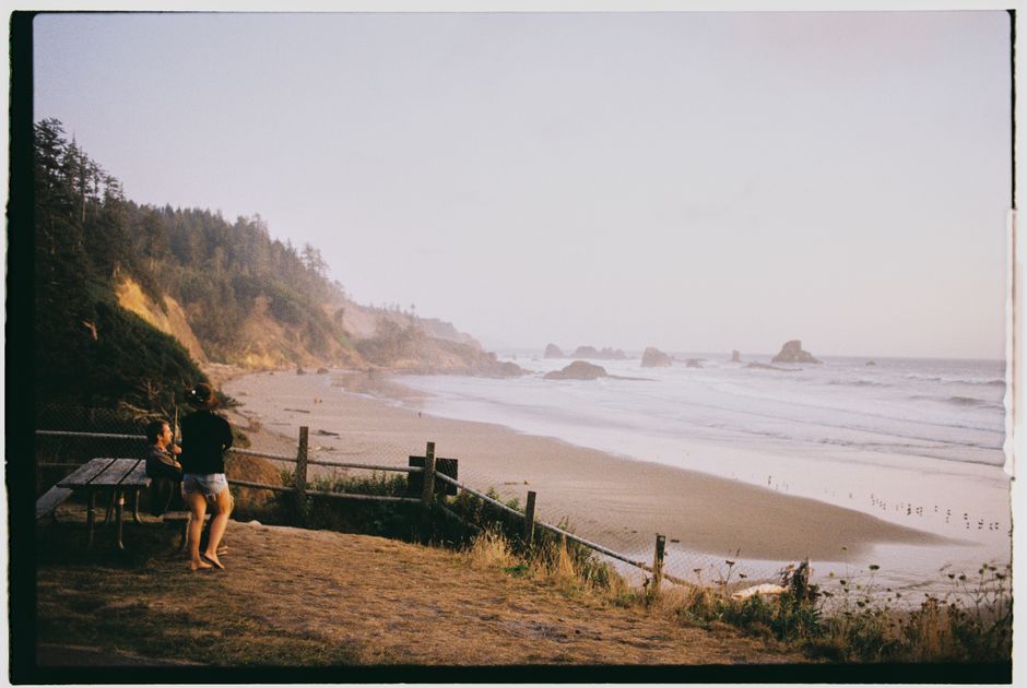 A serene beach with waves, cliffs, and people by a picnic table, captured in a warm light.