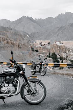 Motorcycles parked by a mountain road, overlooking distant houses and rugged peaks.