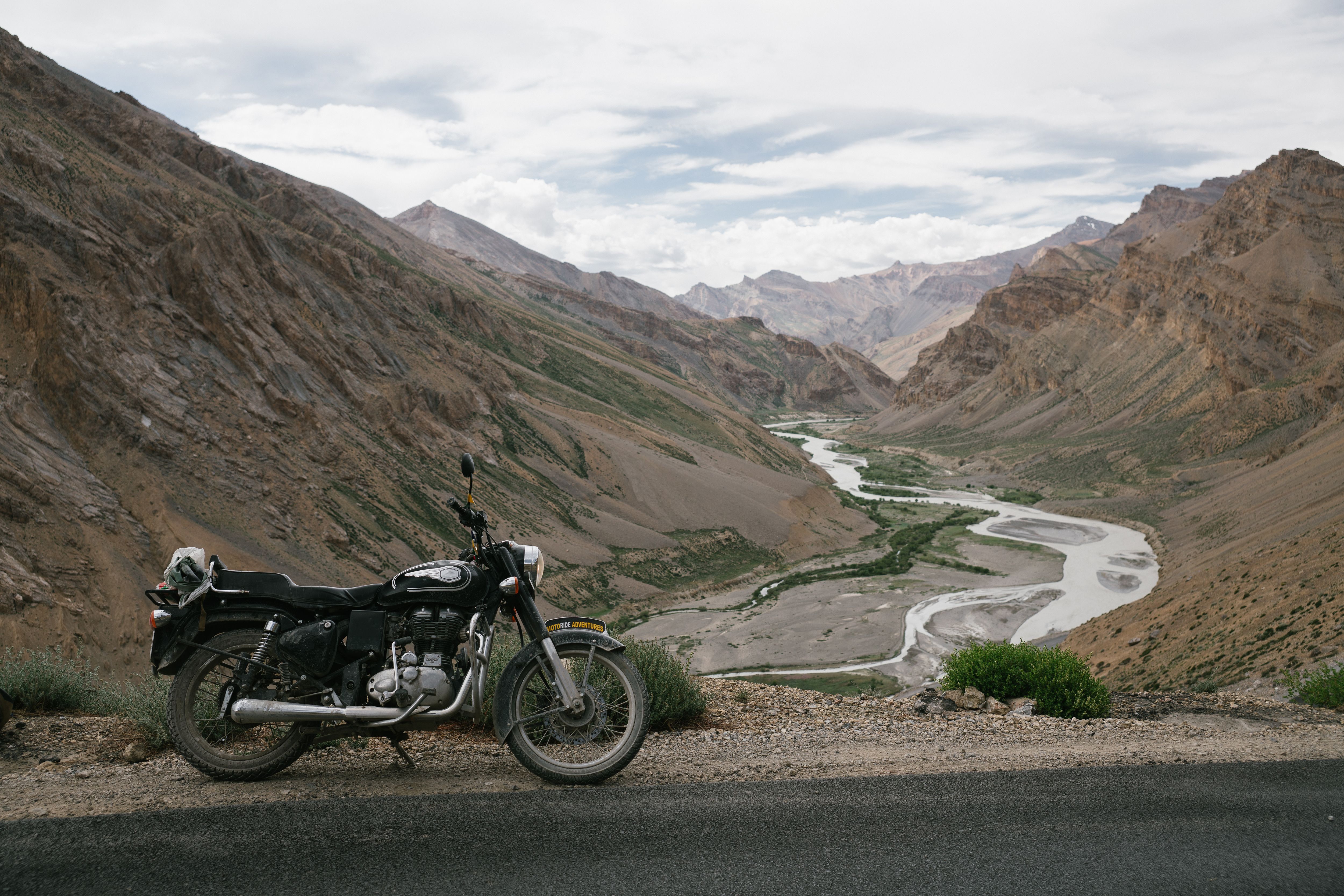 A motorcycle parked along a scenic road with a river winding through mountainous terrain, perfect for adventure trips.