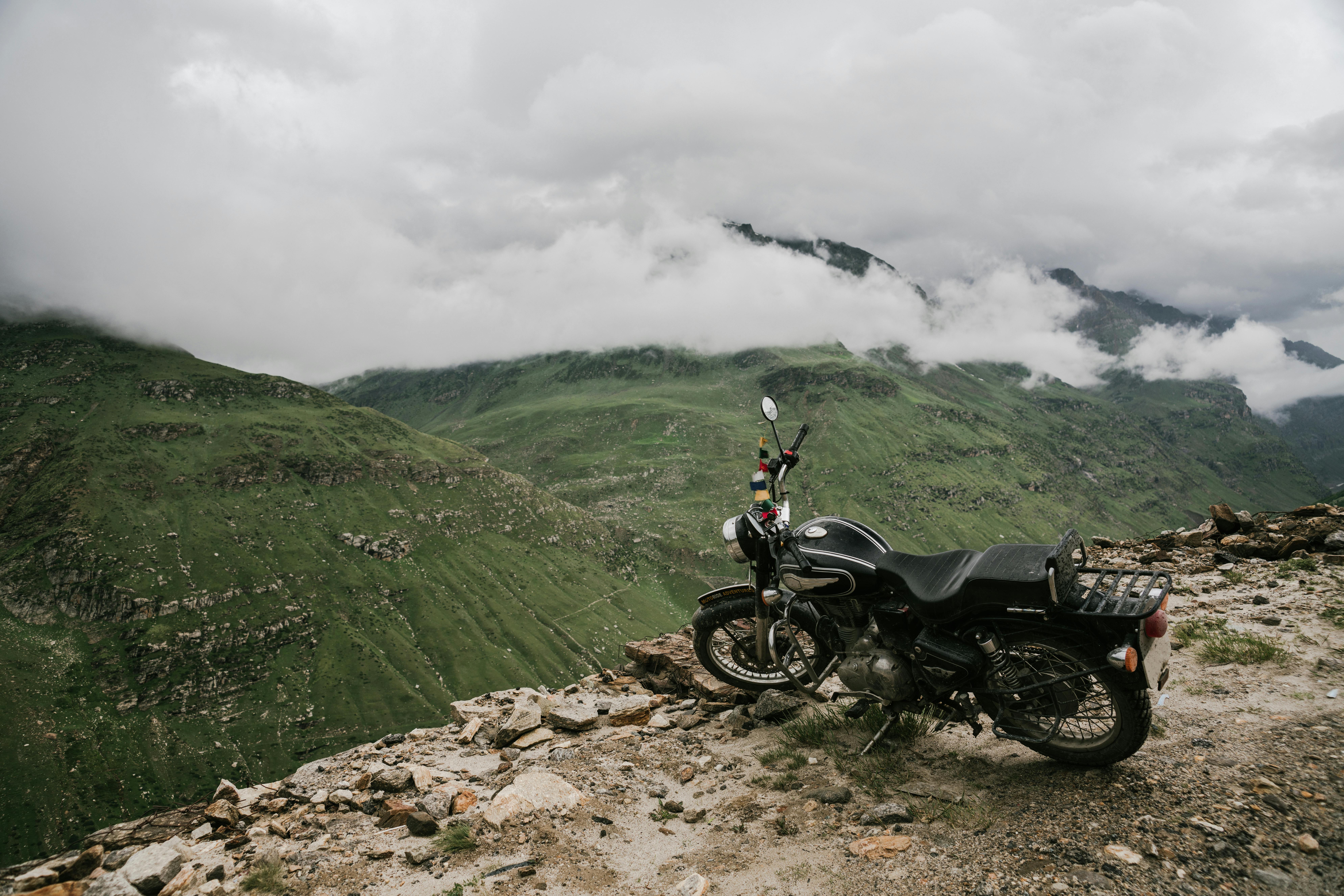 Uma motocicleta estacionada em um penhasco de montanha rochosa com uma vista cênica de montanhas verdes exuberantes e nuvens.